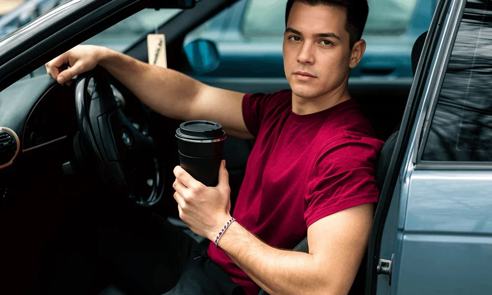 Man in red t-shirt holding coffee cup while sitting in car, hand on steering wheel