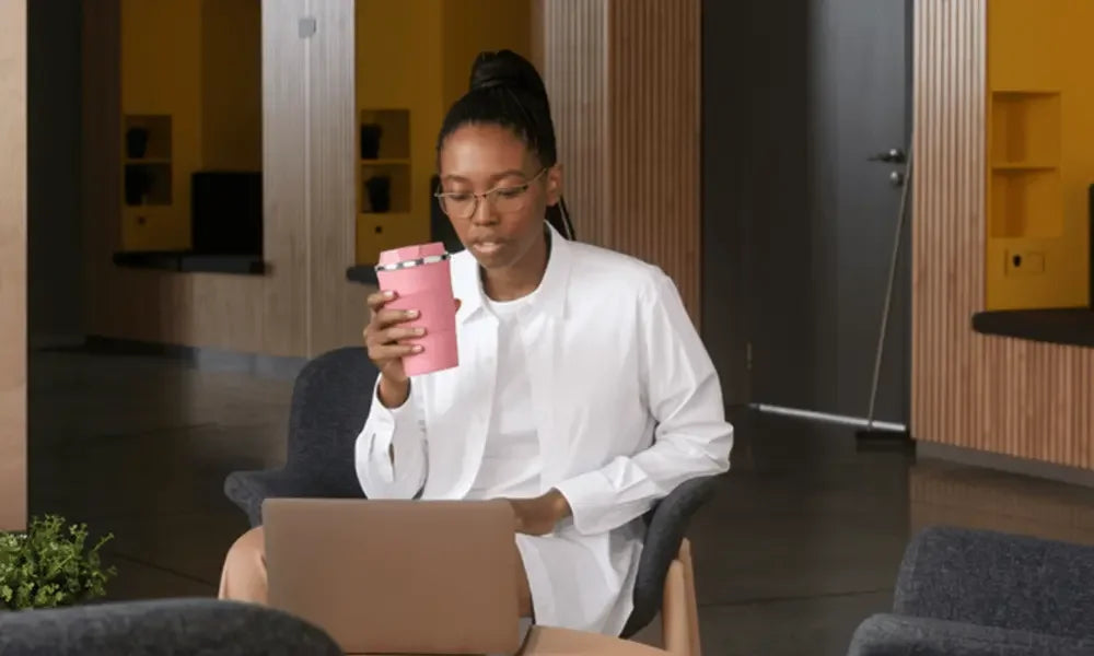 Woman holding pink travel mug while working on laptop in modern office