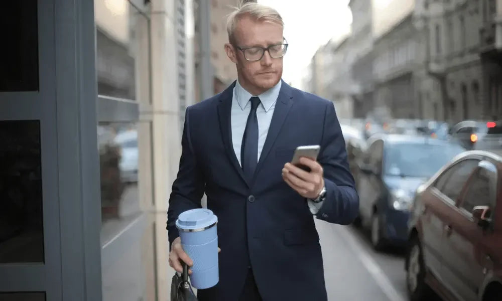 Businessman in suit walking city street, holding phone and blue travel coffee cup, urban commute scene